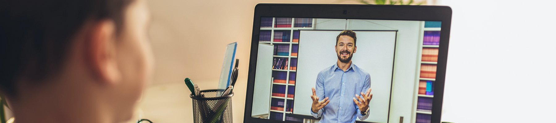 Student attending an online class, watching a virtual lesson with a teacher on a laptop screen.